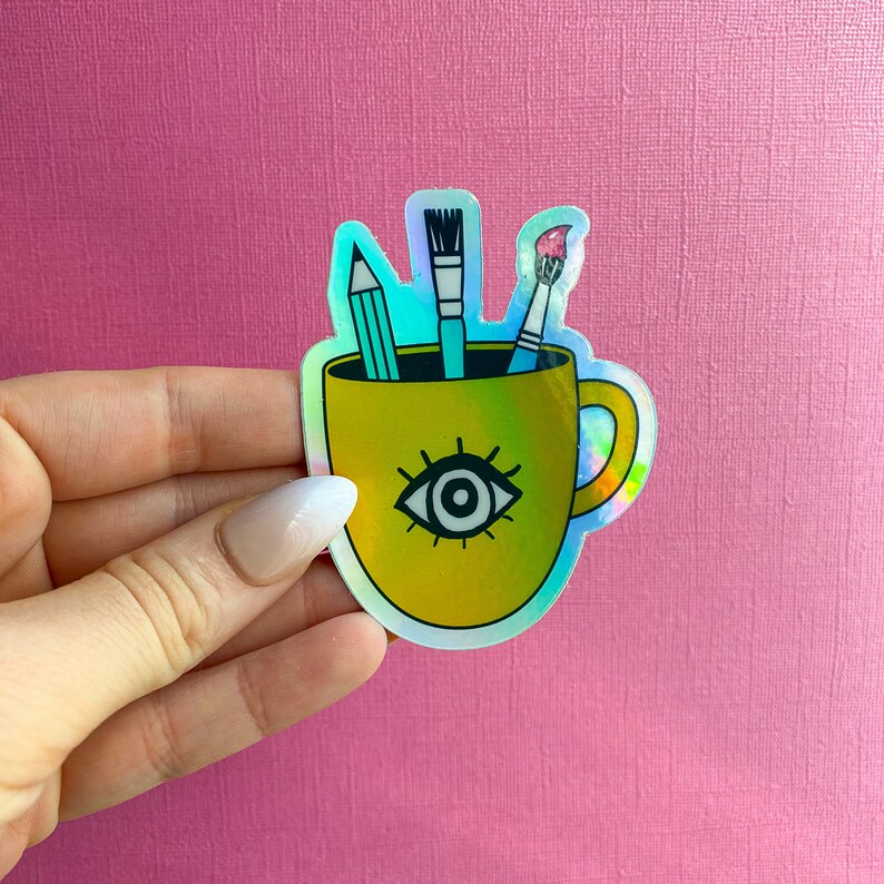 Puede incluir: Una taza de caf&eacute; amarilla con un dise&ntilde;o de un solo ojo y tres pinceles en su interior. La taza tiene un asa y est&aacute; decorada con un acabado hologr&aacute;fico.