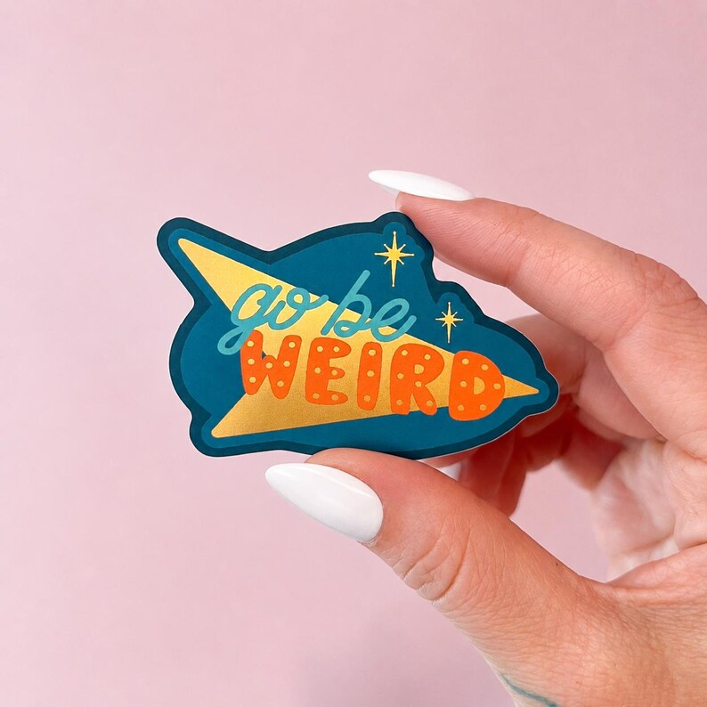 Puede incluir: Una pegatina de color turquesa con forma de cohete retro. El cohete tiene un dise&ntilde;o dorado y naranja. Las palabras "go be WEIRD" est&aacute;n escritas en una fuente l&uacute;dica. Dos estrellas doradas est&aacute;n en el lado derecho de la pegatina.
