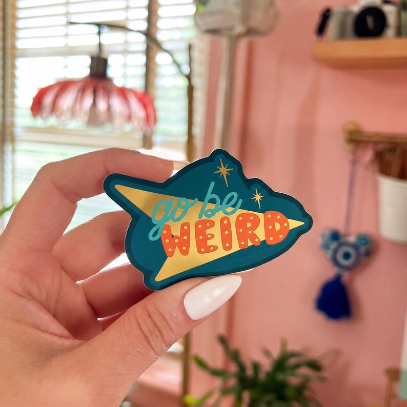 Puede incluir: Una pegatina de color turquesa con un dise&ntilde;o de flecha dorada y las palabras "go be WEIRD" en naranja y blanco. La pegatina tiene una est&eacute;tica retro, de la era espacial. La pegatina se sostiene en una mano.