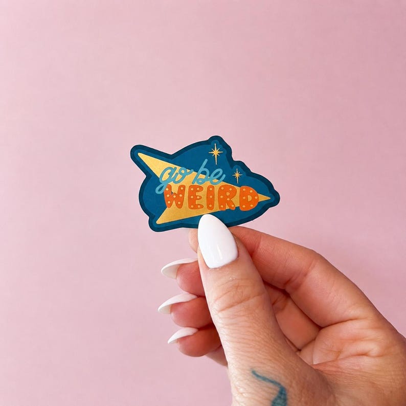 Puede incluir: Una pegatina con las palabras "go be weird" en escritura naranja y azul. La pegatina tiene forma de cohete con un dise&ntilde;o amarillo y azul. El fondo es de color rosa liso.