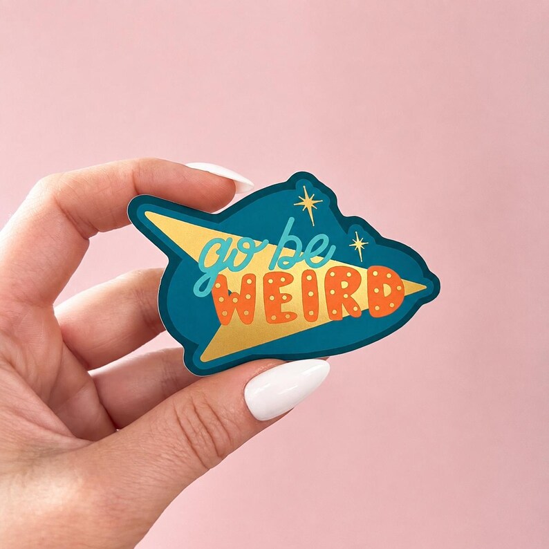 Puede incluir: Una pegatina turquesa con detalles dorados y las palabras "go be WEIRD" en una fuente retro. La pegatina tiene forma de flecha dorada y dos estrellas. El fondo es de color rosa liso.