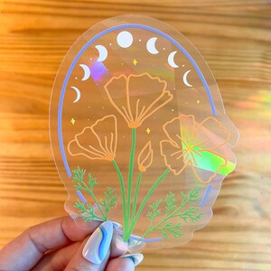 May include: A holographic sticker featuring a cluster of orange poppy flowers with green stems and leaves. The flowers are surrounded by a blue circle with a crescent moon design.
