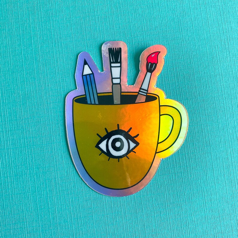 Puede incluir: Una taza de caf&eacute; amarilla con un dise&ntilde;o de un solo ojo y un pincel, un l&aacute;piz y un pincel que sobresalen de la parte superior.