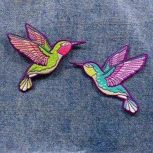 May include: Two embroidered hummingbird patches, one green and pink, the other blue and purple, on a denim background.