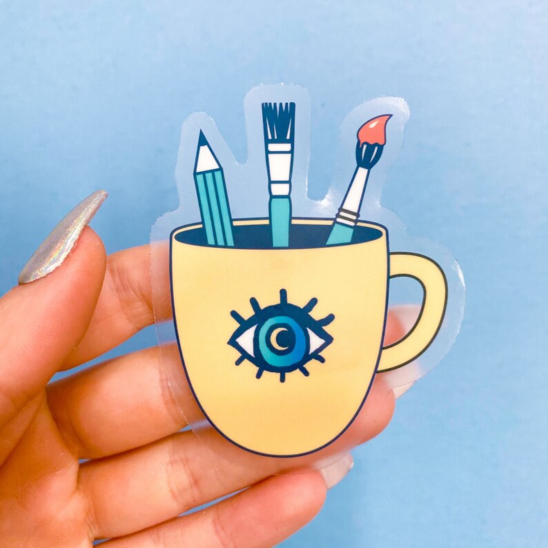 Puede incluir: Una taza amarilla con un dise&ntilde;o de ojo azul y tres &uacute;tiles de arte que sobresalen de la parte superior. Los &uacute;tiles de arte son un l&aacute;piz, un pincel y un pincel.