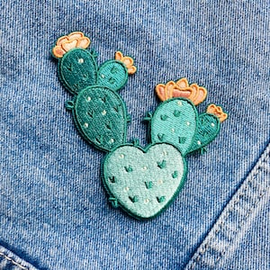 Könnte beinhalten: Gestickter Kaktus-Aufnäher in Türkis und Grün, mit pfirsichfarbenen Blütenakzenten. Der herzförmige Aufnäher hat ein detailliertes Stichdesign, vor einem blauen Jeans-Hintergrund.