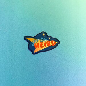 Puede incluir: Una pegatina con un dise&ntilde;o retro y las palabras "go be weird". La pegatina tiene un fondo azul oscuro con una forma de flecha amarilla y texto rojo. Dos peque&ntilde;as estrellas est&aacute;n encima del texto.