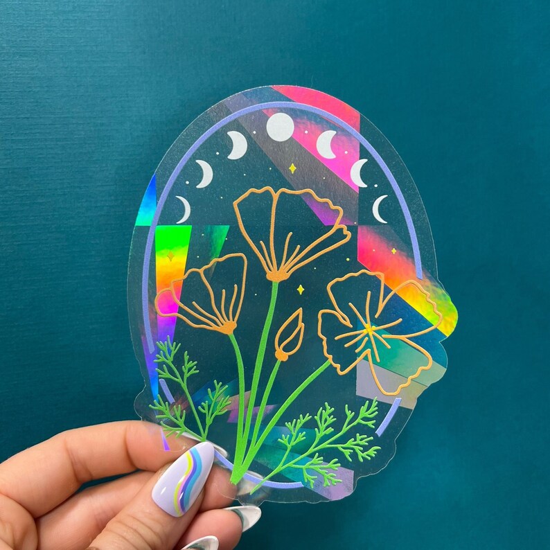 May include: A holographic sticker featuring a floral design with three flowers and leaves. The flowers are outlined in gold and the background is a rainbow gradient with a crescent moon pattern.