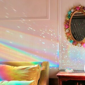 K&ouml;nnte beinhalten: Ein Regenbogen-Prisma-Lichtprojektor wirft farbenfrohe Lichtmuster an eine wei&szlig;e Wand, eine Couch und einen Beistelltisch mit einer Vase mit rosa Blumen. Die Vase ist in einem blaugr&uuml;nen Farbton gehalten und der Spiegel &uuml;ber dem Tisch ist mit bunten Blumen verziert.