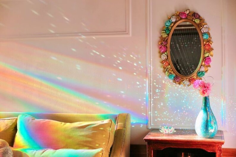 K&ouml;nnte beinhalten: Ein Regenbogen-Prisma-Lichtprojektor wirft farbenfrohe Lichtmuster an eine wei&szlig;e Wand, eine Couch und einen Beistelltisch mit einer Vase mit rosa Blumen. Die Vase ist in einem blaugr&uuml;nen Farbton gehalten und der Spiegel &uuml;ber dem Tisch ist mit bunten Blumen verziert.