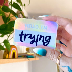 以下が含まれることがあります： 黒い筆記体で「HELLO, I'M trying」と書かれたホログラフィックステッカーです。