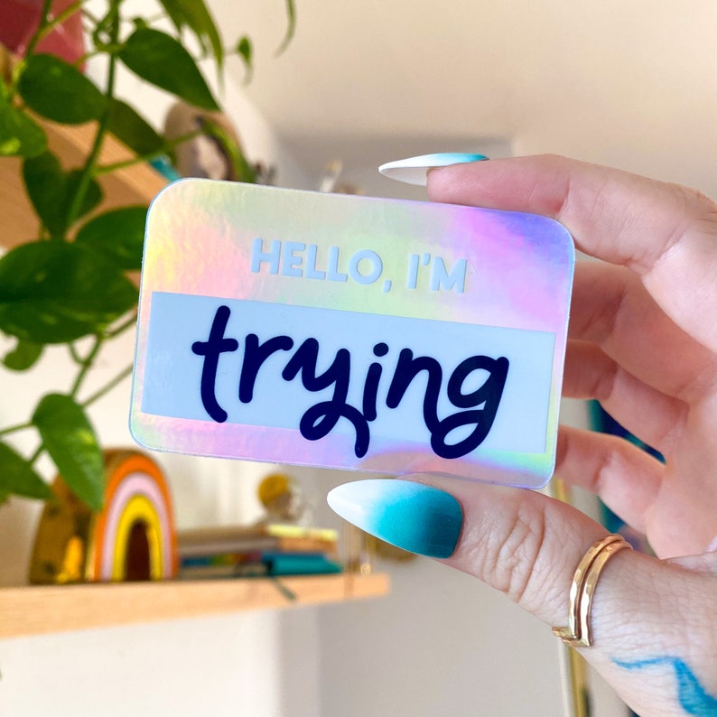 以下が含まれることがあります： 黒い筆記体で「HELLO, I'M trying」と書かれたホログラフィックステッカーです。