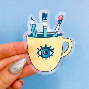 Puede incluir: Una taza de cer&aacute;mica amarilla con un dise&ntilde;o de ojo azul y tres pinceles en su interior. La taza tiene un asa y est&aacute; llena de pinceles.