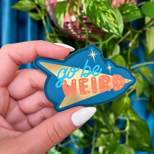 Puede incluir: Una pegatina azul con detalles dorados y las palabras "go be WEIRD" en una fuente retro. La pegatina tiene un dise&ntilde;o de estrella y la sostiene una persona con esmalte de u&ntilde;as blanco.