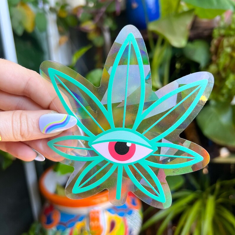 K&ouml;nnte beinhalten: Ein holografischer Aufkleber mit einem gr&uuml;nen Marihuanablatt und einem roten Auge in der Mitte. Der Aufkleber ist schimmernd und hat eine gl&auml;nzende Oberfl&auml;che.