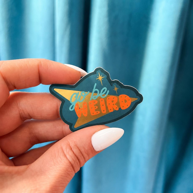 Puede incluir: Una pegatina de color turquesa con las palabras "go be WEIRD" en naranja y amarillo. La pegatina tiene detalles de tri&aacute;ngulo y estrella dorados. La pegatina se sostiene en una mano sobre un fondo azul.