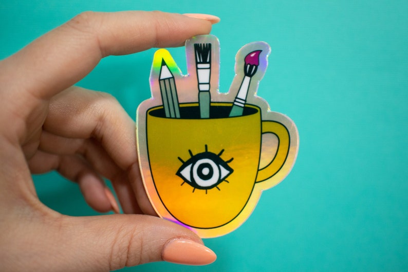 Puede incluir: Una taza amarilla con un dise&ntilde;o de un solo ojo y un acabado hologr&aacute;fico. La taza tiene un asa y tres art&iacute;culos de arte en su interior: un l&aacute;piz, un pincel y un pincel con pintura rosa.