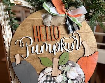 May include: A wooden round sign with the text "Hello Pumpkin" in white letters. The sign is decorated with three pumpkins, one in orange, one in black and white checkered pattern, and one in gray. The sign is also decorated with a green and white bow.