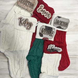 May include: Five white, red and green knitted Christmas stockings with personalised wooden name tags. The names on the tags are Kiley, Houston, Emma, Rome and Jackson.