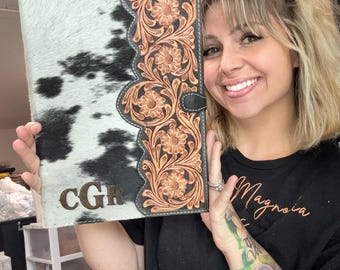 Puede incluir: Una carpeta de cuero de vaca blanco y negro con un diseño de cuero estampado en marrón en el lado derecho. La carpeta tiene las iniciales "CGR" grabadas en la parte delantera.