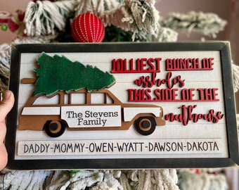 May include: A wooden sign with a black frame and white background. The sign features a cutout of a station wagon with a Christmas tree on top. The text on the sign reads "JOLLIEST BUNCH OF ASSHOLES THIS SIDE OF THE NUTHOUSE" and "The Stevens Family" and "DADDY-MOMMY-OWEN-WYATT-DAWSON-DAKOTA".