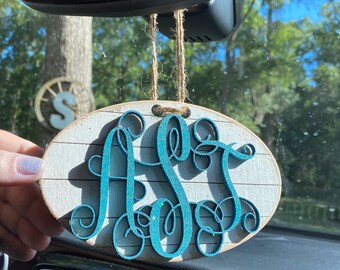May include: A teal blue monogrammed car air freshener with the letters "ASS" in a cursive font. The air freshener is made of wood and has a white and gray striped background. It is hanging from a rearview mirror in a car.