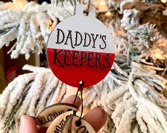 May include: A white and red wooden Christmas ornament with the text "Daddy's Keepers" on the front. Three wooden fish ornaments with the names "Owen", "Liam", and "Noah" are hanging from the ornament.