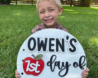 May include: A white round sign with the text "Owen's 1st day of Second Grade" in black lettering. A red apple with a green stem and leaf is next to the text "1st". A pink and yellow pencil is below the text. A young boy is holding the sign in front of him.