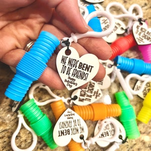 Op de afbeelding: Een verzameling kleurrijke, buigzame plastic buizen met bevestigde hartvormige houten labels. De labels hebben de tekst "We were bent to be friends!" en "Happy Valentine's Day".