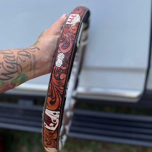 Puede incluir: Una funda circular para volante de cuero marrón con un intrincado grabado floral en marrón oscuro. El diseño incluye detalles en blanco y rojo que representan naipes, un par de dados y un emblema de escudo rojo con el número 777 en blanco.