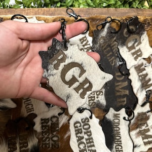 Op de afbeelding: Een verzameling koeienhuid sleutelhangers met verschillende gegraveerde namen. De sleutelhangers zijn bruin, zwart en wit met een koeienhuidpatroon. De namen op de sleutelhangers zijn: R.G., Sophia Grace, McDonough, Chloe Reese, Hugh Tree en Wyatt.