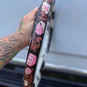 Op de afbeelding: Een zwarte stuurwielhoes met een bruin, bloemrijk lederen patroon en roze grafische accenten, waaronder een roze gokkast-icoon met het getal 777, roze dobbelstenen en roze pokerfiches, afgewerkt met witte contraststiksels.