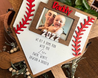 May include: Wooden baseball bat picture frame with a red and white baseball stitching design. The frame has a photo of a father and son with the text "Daddy" above the photo. The text below the photo reads "We hit a home run with you. Love Owen, Wyatt, Dawson & Dakota."
