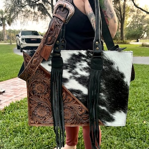 Puede incluir: Un bolso tote de piel de vaca blanco y negro con una correa de cuero marrón y detalles de flecos. La correa tiene un intrincado trabajo de estampado floral.