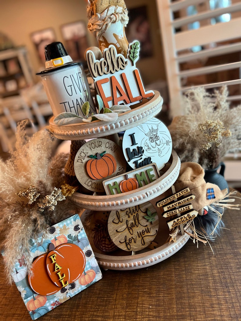 May include: A three-tiered white tiered tray decorated with fall-themed wooden signs, a white mug with black lettering that says 'Give Thanks', a brown and white pumpkin, and a brown and white scarecrow. The signs include 'hello fall', 'I love fall', 'Pick your own local', 'Home', 'I still fall for you', and 'Pumpkin Patch', 'Hayrides', 'Corn Maze', and 'Apple Orchard'. The tray is on a brown wooden surface.