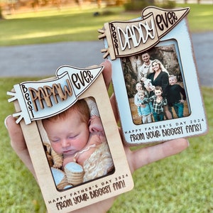 May include: Two wooden picture frames with a white background and a banner at the top. The banner on the left frame says "Best Papaw Ever" and the banner on the right frame says "Best Daddy Ever". Both frames have a message at the bottom that says "Happy Father's Day 2024 from your biggest fan!"