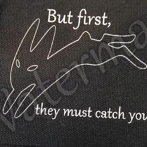 Peut inclure: Écusson carré noir avec texte blanc et illustration de lapin. Le texte dit « Mais d'abord, ils doivent vous attraper. » L'écusson a un bord brut et est placé sur une surface bleue. Une règle est en bas.