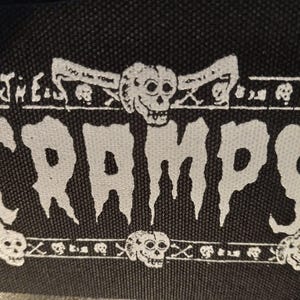 May include: Black patch with the white text "THE CRAMPS" in a dripping font. The patch has a border with skulls and crossbones. The top border has the text "DO THE YOW".