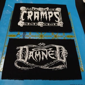 May include: Two black fabric patches with white text. The top patch reads "THE CRAMPS" with skull and crossbones imagery. The bottom patch reads "THE DAMNED" with a stylized design. Both patches are rectangular.