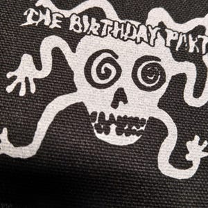 May include: Black fabric with a white graphic of a skull with spiral eyes and the text "THE BIRTHDAY PARTY" above it. The skull has a wide grin and stylized limbs extending from the sides.