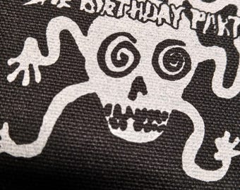 Puede incluir: Tela negra con un gráfico blanco de una calavera con ojos en espiral y el texto "THE BIRTHDAY PARTY" encima. La calavera tiene una amplia sonrisa y extremidades estilizadas.