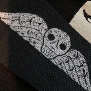 May include: Close-up of black fabric featuring a white skull and wing design. The skull has detailed eye sockets and teeth, and the wings are spread out. The design is likely screen printed.