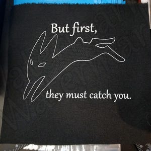 Peut inclure: Carré de tissu noir avec texte blanc et un dessin au trait d'un lapin. Le texte se lit "But first, they must catch you."