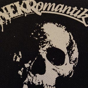 May include: Black patch featuring a white skull graphic and the word "NEKRomantik" in a stylized font. The design has a distressed, grunge aesthetic. The patch is likely for clothing or accessories.