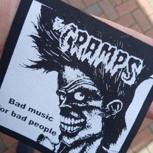 May include: A black and white patch featuring the band name "CRAMPS" in a bold, stylized font. Below the band name is a graphic of a zombie-like face. The text "Bad music for bad people" is printed below the graphic.