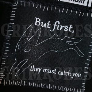 Peut inclure: Écusson carré en tissu noir avec couture et texte blancs. L'écusson présente l'expression "But first," au-dessus d'une illustration de lapin blanc, et les mots "they must catch you" en dessous. Le patch a des coins arrondis.