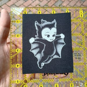 May include: A black square patch featuring a cartoon bat-like character with a smiling face and outstretched wings. The character is in white, with black outlines, on a black background. The patch is being measured with a yellow ruler.