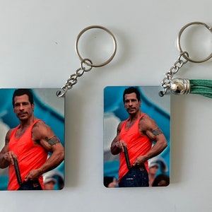 May include: Two keychains with a photo of a man wearing a red tank top and blue jeans. The keychains are silver and one has a green tassel.