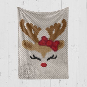 May include: A crocheted blanket featuring a reindeer face with brown antlers, a red bow, black eyelashes, and a red nose.