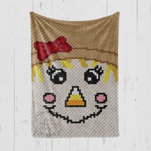 May include: A crochet blanket featuring a cute cartoon scarecrow face with a straw hat, red bow, and candy corn nose.